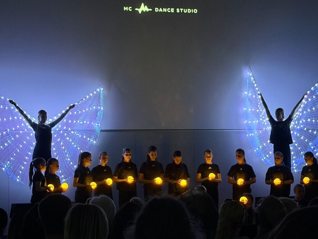 Zwei Tänzerinnen mit leuchtenden Flügeln, davor eine Reihe Kinder mit Lichtkugeln